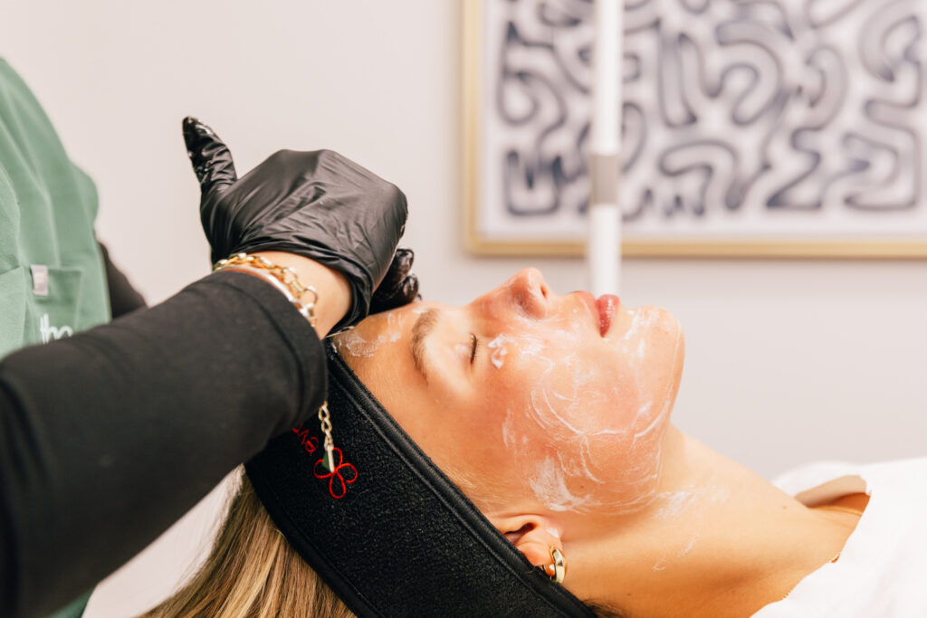 A patient lies on her back as a chemical peel near Lenexa is applied to her face. Her provider is wearing surgical gloves as they spread the peeling solution on her cheeks and forehead.