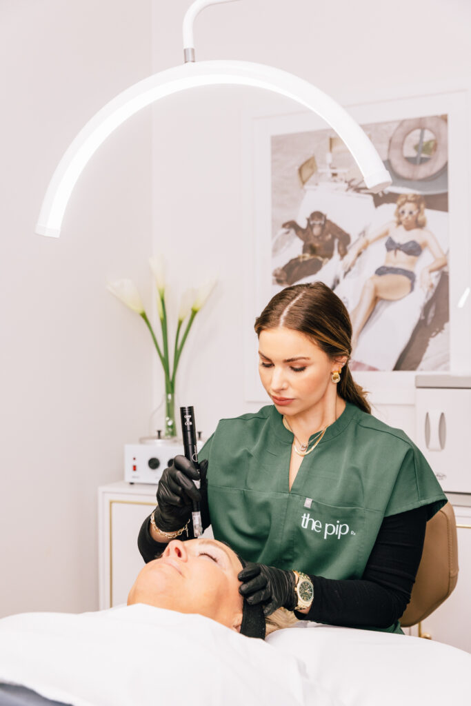 A provider at The Pip Rx administers a microneedling treatment in Overland Park. Wearing surgical gloves, she carefully passes the handheld microneedling device over her patient's forehead.